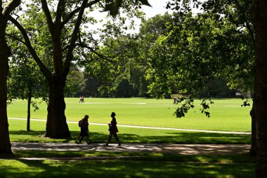 Londra, İngiltere - Haziran 2021: Londra şehir merkezindeki Hyde Park 'ta yürüyen insanların silueti. Manzara ağaçlar tarafından çerçevelenir.