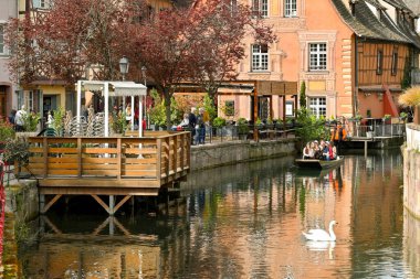 Colmar, Fransa - Nisan 2022: Kanaldaki küçük bir teknede turistler, önlerinde bir kuğuyla birlikte şehir boyunca ilerliyorlar.