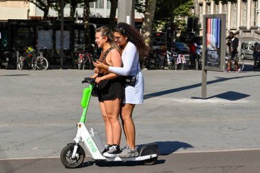 Antwerp, Belgium - August 2022: Two people balancing on an electric scooter in the c ity centre. One of the people is using a mobile phone while holding on.