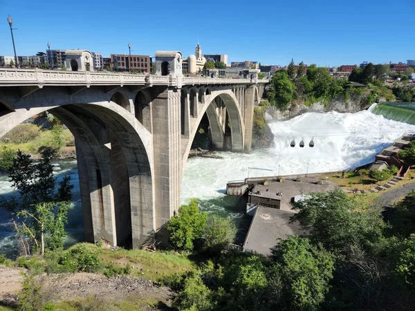 Torrential flow over Lower Spokane Falls under Monroe Street Bridge in downtown Spokane after heavy rains on bright sunny summer morning.