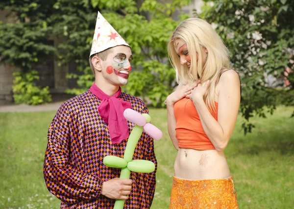 Portrait of a smiling man dressed like a clown trying to present a ballon to a happy girl with butterfly makeup — Stock Photo, Image