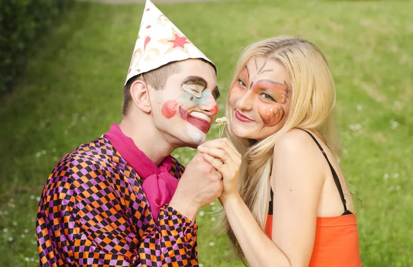 Portrait of a smiling man dressed like a clown trying to present a flower to a happy girl with butterfly makeup Royalty Free Stock Photos