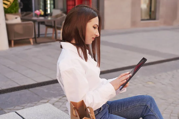 Genç ve güzel bir kadın şehirde yürüyor. Sokakta elinde telefon ve tabletle dolaşan bir kızın portresi. Paket almak ve alışveriş yapmak. Genç ve güzel bir kadın şehirde yürüyor. Çevrimiçi iş, uzaklık öğrenimi