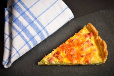 Soğanlı, yumurtalı ve pastırmalı bir dilim turta. Geleneksel Fransız kiş Lorraine. Pişmiş krema hamuru. Siyah arka planda tekstil mutfak havlusuyla taş tabağa dilimlenmiş turtayı kapat