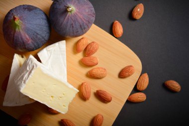 Tahta tahtadaki yumuşak Fransız Brie peyniri parçalarının üst görüntüsü koyu arka planda badem fıstıklarıyla servis ediliyor. Peynir tahtası. Arkaplanda incir meyveleri olan tabaklar
