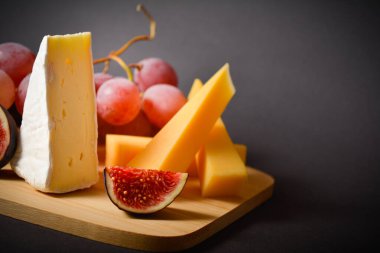 Sert ve yumuşak peynirli peynir tahtası, kırmızı üzüm ve incir dilimleri. Siyah arka planda bir tabak. Beyaz küflü Fransız peyniri. Yatay görüntü. Aperatifler ve başlangıçlar. Karanlık ruh hali fotoğraf tarzı.