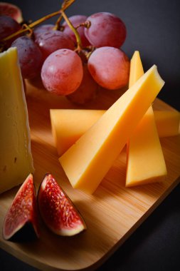 Koyu arkaplanda meyve servis edilen peynir tahtası. Üzüm ve incir dilimli sert ve yumuşak Fransız peyniri parçaları. Tabakta beyaz küflü peynir. Tahta tahtada aperatifler. Meyveler ve meyveler