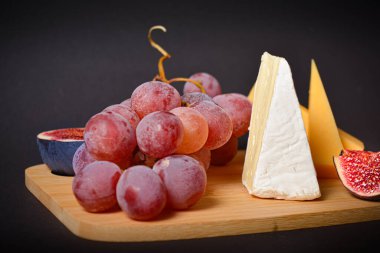 Koyu arkaplanda servis edilen peynirin yan görüntüsü. Üzüm ve incir dilimli sert ve yumuşak Fransız peyniri parçaları. Tabakta beyaz küflü peynir. Tahta tahtada aperatifler. Meyveler ve meyveler