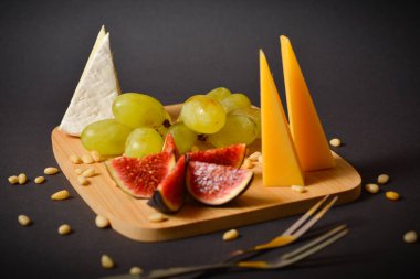 Çam fıstıklı sert ve yumuşak peynirli peynir tabağı, taş tabakta servis edilen incir dilimleri ve üzüm. Siyah arka planda iki meyve çatalı. Fransız Brie peyniri tepecikli peynirin yan görünümü.