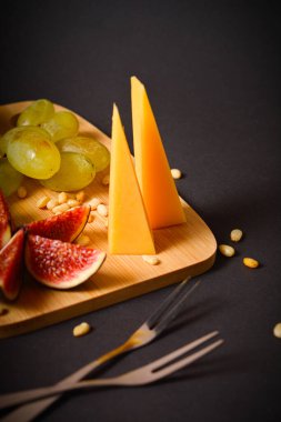 Siyah arka planda çam fıstığı olan peynirin yan görüntüsü. Yeşil üzüm ve incir dilimli sert Fransız peyniri parçaları. Peynir tabağı. Tahta tahtada aperatifler. Tabağın yanında iki meyve çatalı var.