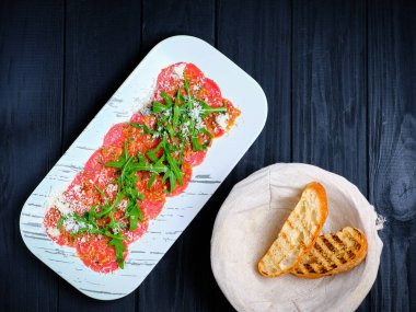 Taze roka yapraklarıyla servis edilen ince sığır eti carpaccio dilimleri ve koyu arkaplanda beyaz tabakta rendelenmiş parmesan peyniri. İtalyan geleneksel yemeğinin yanında iki ızgara ciabatta ekmeği dilimi. Çiğ et.