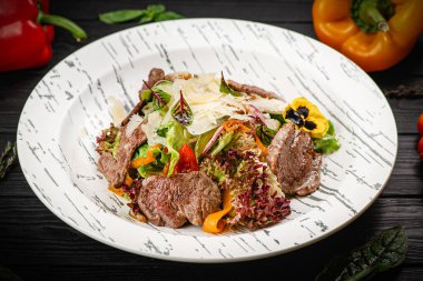 Siyah ahşap arka planda beyaz tabakta servis edilen sıcak sığır eti salatası. Kızarmış dana madalyonu, kabak dilimleri, kiraz domatesleri, parmesan peyniri ile servis edilen marul. Arka planda sebzeler