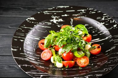Klasik İtalyan Caprese tarzı salata, vişneli domates, mini bebek Mozzarella peyniri, taze roka ve fesleğen yaprağı pesto ve balzamik sosla servis edilir. Siyah tabakta sağlıklı salata, ahşap arka plan