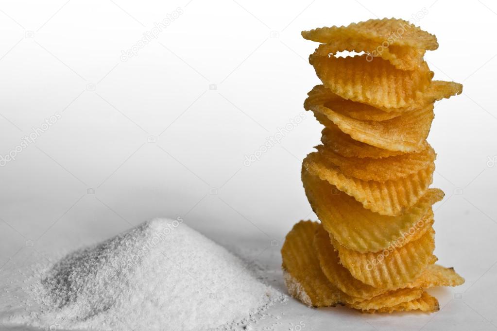Stack of wavy chips placed next to piles of salt, on the white background