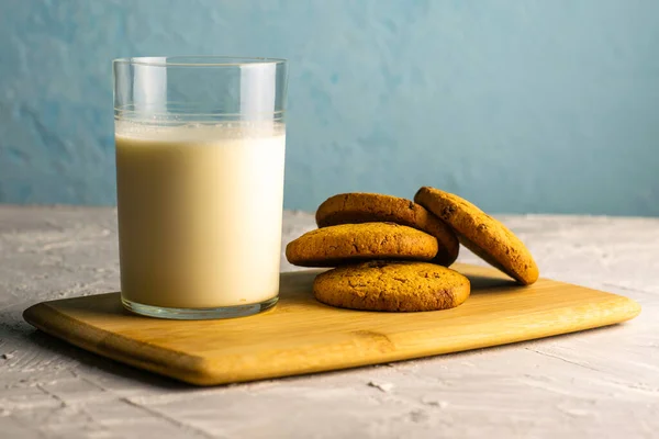 Masadaki tahta kesme tahtasının üzerinde yulaf ezmeli kurabiye ve bir bardak süt. — Stok fotoğraf