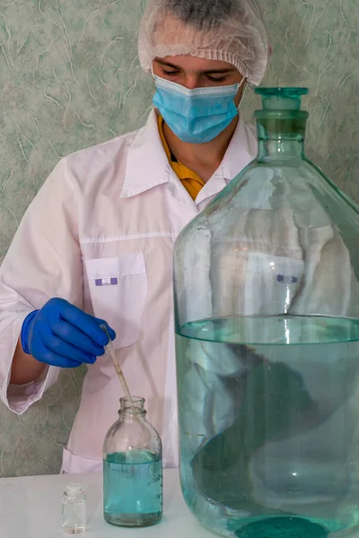 Лабораторний асистент у білих лабораторних рукавичках для захисної маски та кепка в лабораторії витягує рідину з бульбашки з піпеткою поруч із судинами інших розмірів — стокове фото