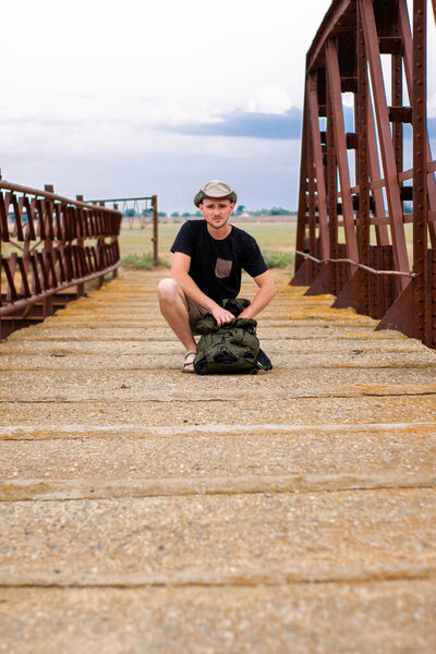 a young tourist guy in a hat with a backpack is sitting on an old iron bridge across the river. High quality photo