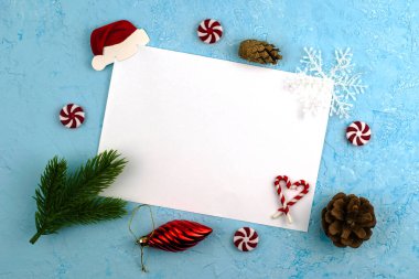 Yeşil köknar dalları, lolipoplar, kozalaklar, Noel süslemeleri ve kar taneleriyle çerçevelenmiş mavi bir yüzeyde beyaz bir yaprak. Noel Baba 'ya yeni yıl dilekleri ile yazılmış kavramsal bir mektup.