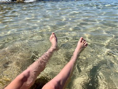 Genç bir çocuk kumlu sahilde suda oturuyor ve yaz gününün tadını çıkarıyor. Çıplak ayaklı çocuk. Deniz kenarında bir tatil. Açık havada..