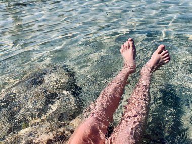 Genç bir çocuk kumlu sahilde suda oturuyor ve yaz gününün tadını çıkarıyor. Çıplak ayaklı çocuk. Deniz kenarında bir tatil. Açık havada..