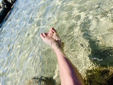 Genç bir çocuk kumlu sahilde suda oturuyor ve yaz gününün tadını çıkarıyor. Çıplak ayaklı çocuk. Deniz kenarında bir tatil. Açık havada..