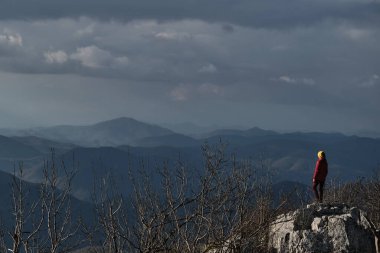 Kırmızı ceketli ve parlak sarı şapkalı gezgin kız uçurumun tepesinde duruyor ve uzaktaki dağların manzarasından hoşlanıyor. Doğa turistlerinin dağlardaki görüntüsü.