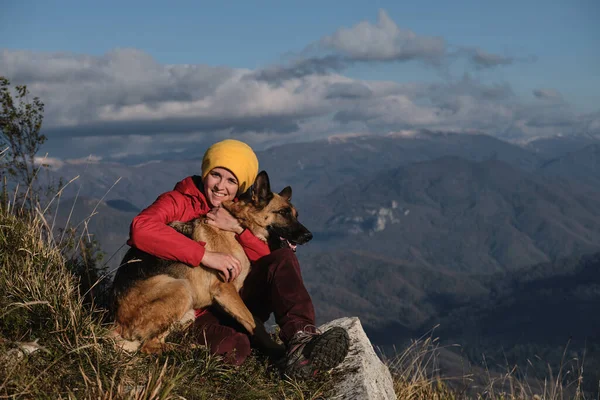 Kırmızı ceketli genç beyaz kadın dağın tepesinde köpeğinin yanında oturuyor ve gün batımında manzaranın tadını çıkarıyor. Alman çoban köpeği ve sahibiyle seyahat et. Doğadaki en iyi arkadaşa sarılmak.
