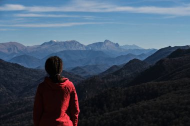 Parlak kırmızı ceketli kadın gezgin arkası dönük duruyor ve ufuktaki karlı dağlara bakıyor. Turist kızın saç stili..