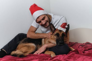 Sakallı ve kırmızı Noel Baba şapkalı genç Avrupalı adam battaniyenin üzerinde oturuyor ve Alman çoban köpeğine sarılıyor. Noel 'i ve yeni yılı evde köpekle birlikte kutlayın..