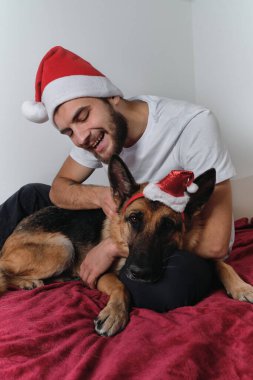 Sakallı ve kırmızı Noel Baba şapkalı genç Avrupalı adam battaniyenin üzerinde oturuyor ve Alman çoban köpeğine sarılıyor. Noel 'i ve yeni yılı evde köpekle birlikte kutlayın..