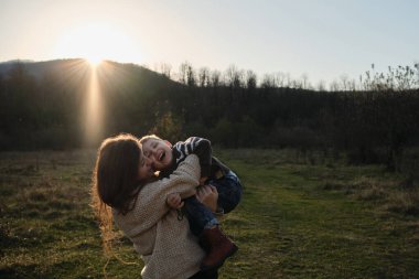 Çizgili kazaklı beyaz çocuk neşeyle kadının kollarında oturuyor ve ikisi de gülüp sarılıyorlar. Çocukluğunu doğada sevgili annenle geçir. Anneliğin neşesi. Sıcak batan güneş.
