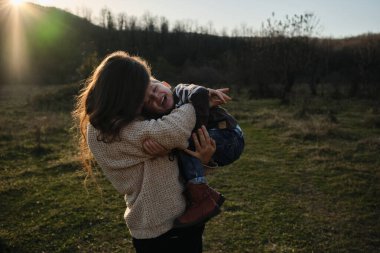 Çizgili kazaklı beyaz çocuk neşeyle kadının kollarında oturuyor ve ikisi de gülüp sarılıyorlar. Çocukluğunu doğada sevgili annenle geçir. Anneliğin neşesi. Sıcak batan güneş.