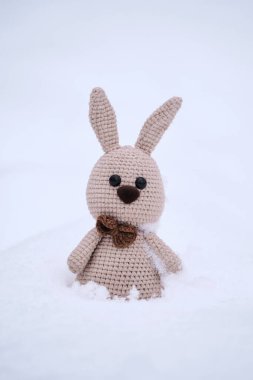 Uzun kulaklı, kahverengi burunlu ve papyonlu bej amigurumi tavşanı. Beyaz kardaki oyuncak tavşan. Paskalya tavşanı. Çocuk için Noel hediyesi el yapımı hayvan oyuncağıdır..