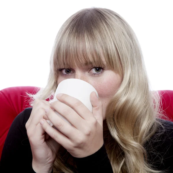 Young blond haired girl drink cup of tee on red sofa in front of — Stock Photo, Image