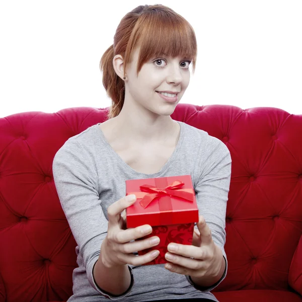 Junge schöne rothaarige Mädchen auf rotem Sofa hat eine Geschenkbox für — Stockfoto