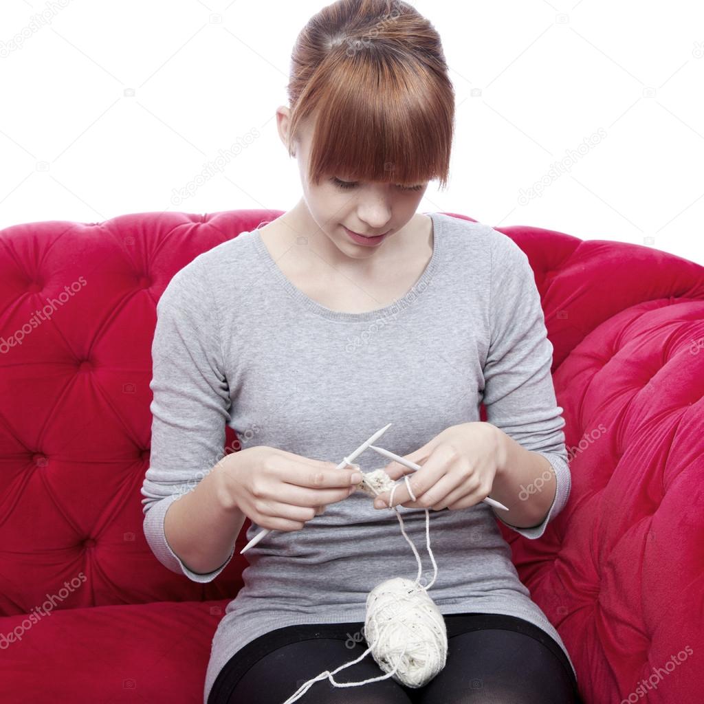 young beautiful red haired girl knitting on red sofa in front of