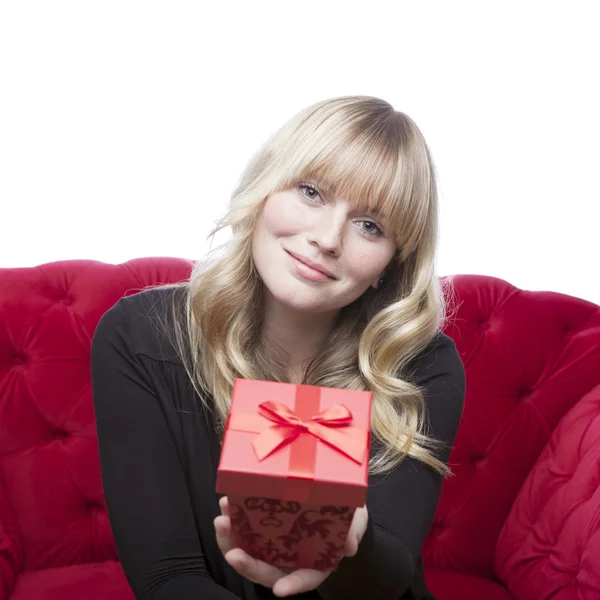 Junge blondhaarige Mädchen hat eine Geschenkbox für Sie auf rotem Sofa in — Stockfoto
