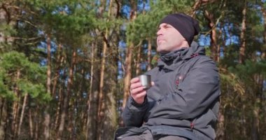 Kışlık giysiler içindeki adam nehir kıyısında bir çam ormanında çay içer ve dinlenir. 