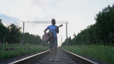 Güneş gözlüklü ve şapkalı bir adam demiryolunda yürüyor ve gitar çalıyor.