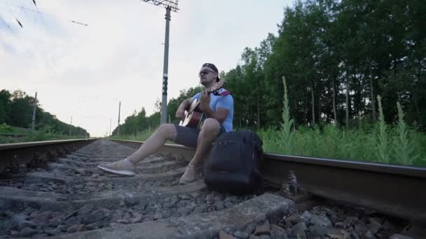 Человек в солнцезащитных очках и кепке сидит на рельсах железной дороги и играет на гитаре — стоковое видео