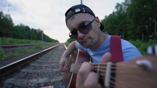 Músico con gafas de sol y gorra sentado en el ferrocarril y toca la guitarra — Vídeos de Stock