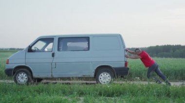 Adam tarlaya doğru yuvarlanmaya başlayan eski bir minivan arabasını arkadan itiyor.
