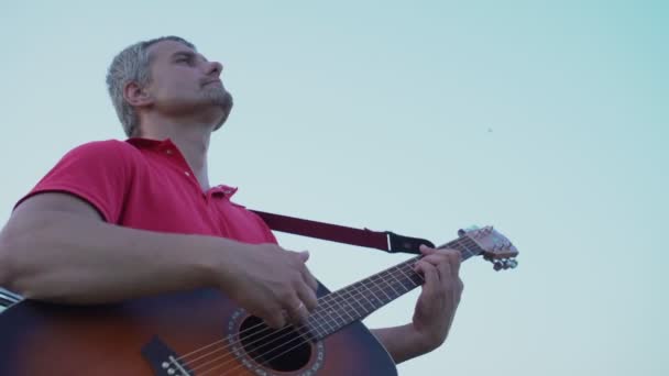 Hombre reflexivo toca la guitarra acústica contra el fondo del cielo azul claro — Vídeos de Stock