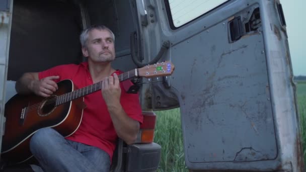 El hombre toca en la guitarra acústica sentado en el borde del cuerpo de la vieja minivan — Vídeo de stock