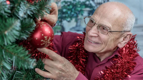 头戴眼镜的老男人把红球挂在圣诞树上. 免版税图库照片