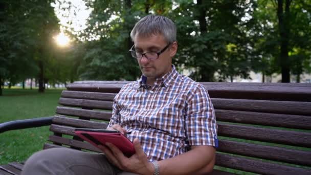 El hombre trabaja duro en la tableta sentado en el banco en el parque de la ciudad — Vídeos de Stock