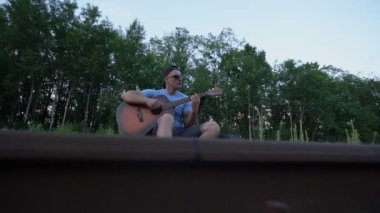 Sırt çantalı bir adam demiryolunda oturmuş gitar çalıyor ve şarkı söylüyor.