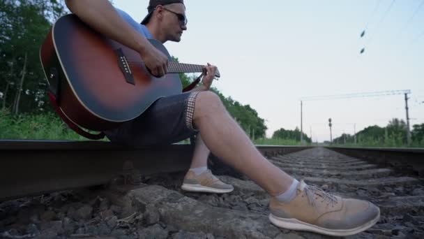 Hombre se sienta sobre rieles de ferrocarril toca la guitarra y mirando hacia atrás de cerca — Vídeos de Stock