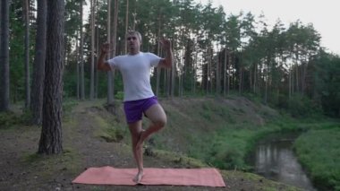 Formda, rahat bir adam, beyaz bluz ve şort giymiş olarak ağacın üstünde duruyor..