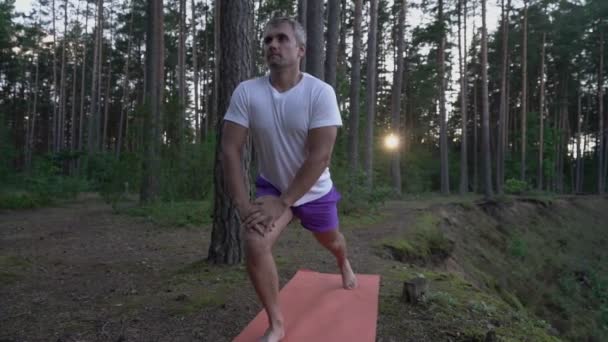 Зрелый человек делает низкую позу выпада на коврике для йоги в лесу. — стоковое видео